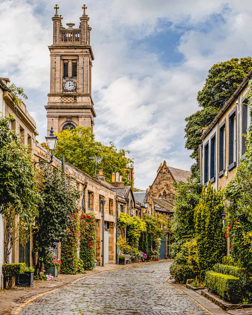 A charming cobblestone street lined with quaint buildings, including a clock tower with a distinctive architectural style, surrounded by lush greenery in the background.
