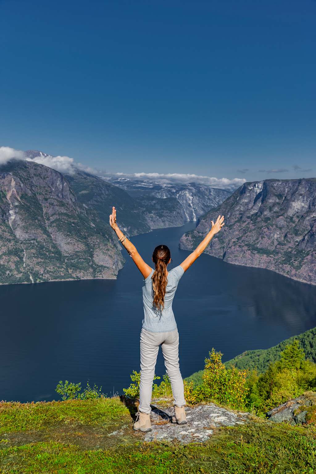 A person with arms raised stands on a grassy hilltop overlooking a vast, scenic lake surrounded by mountains under a clear blue sky.