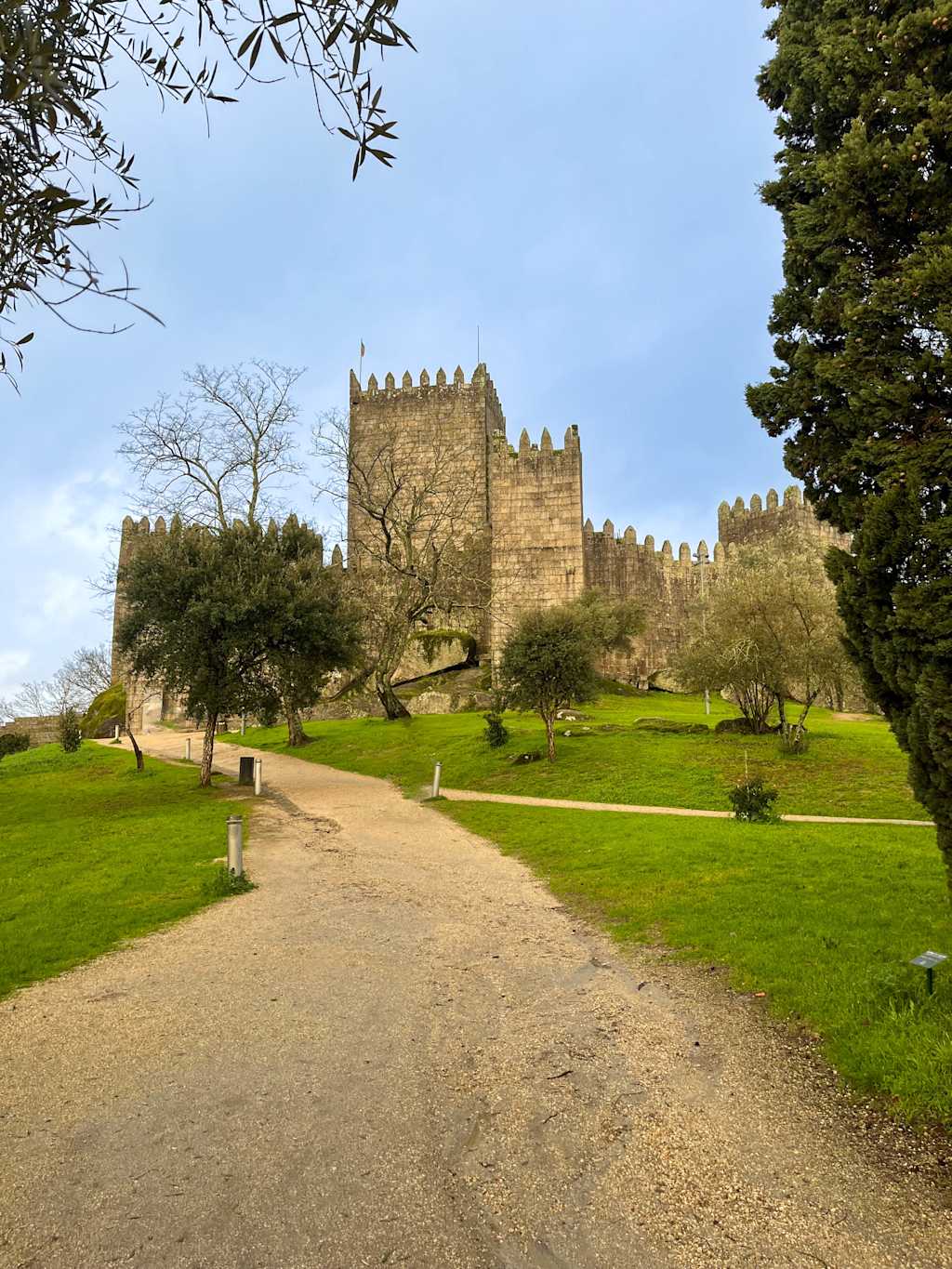 A winding gravel path leads through a lush, grassy landscape towards a grand, medieval-style castle with ornate towers and battlements, surrounded by trees and set against a clear blue sky.