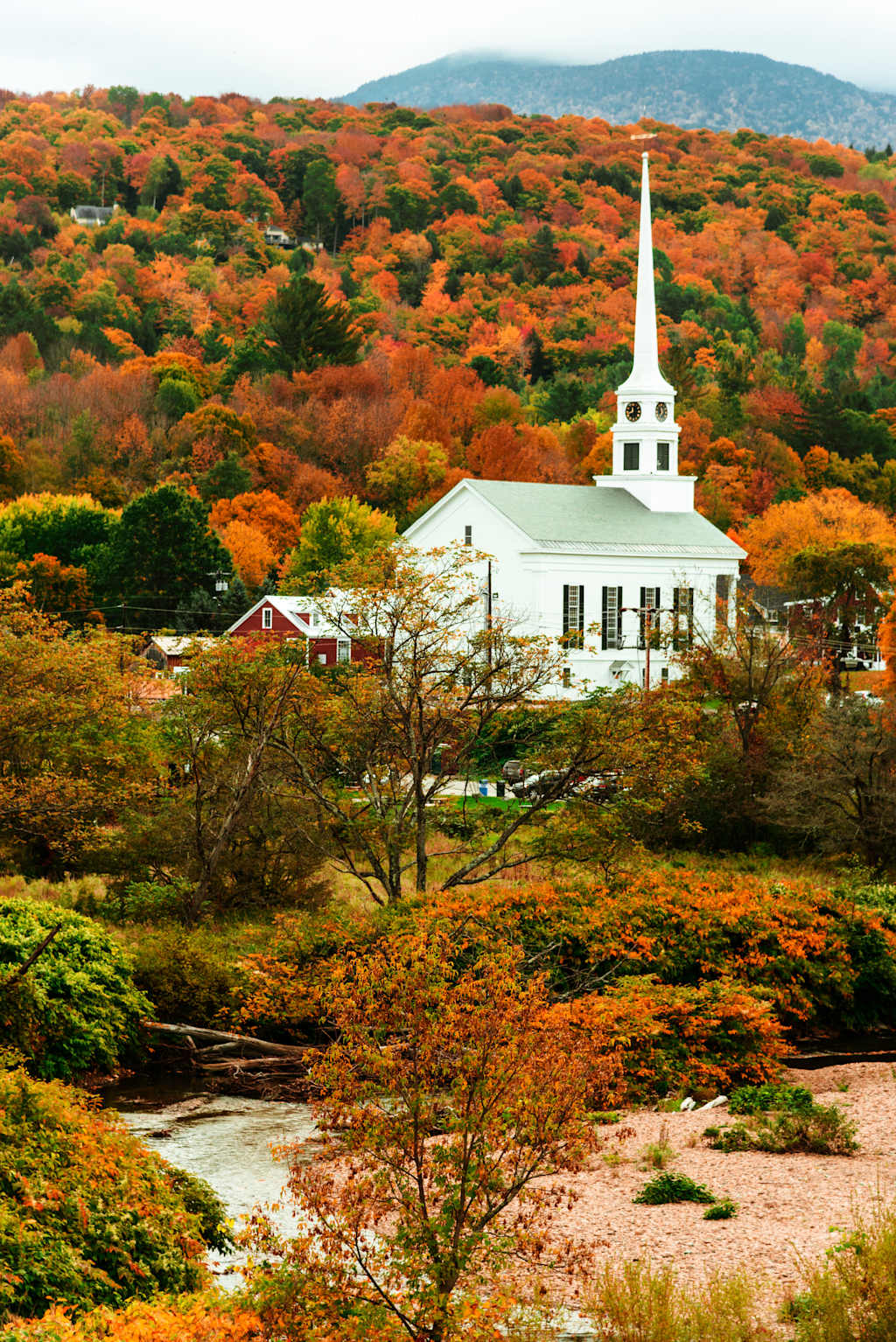 A picturesque New England town nestled amidst a vibrant autumnal landscape, with a white church steeple rising above the colorful foliage.