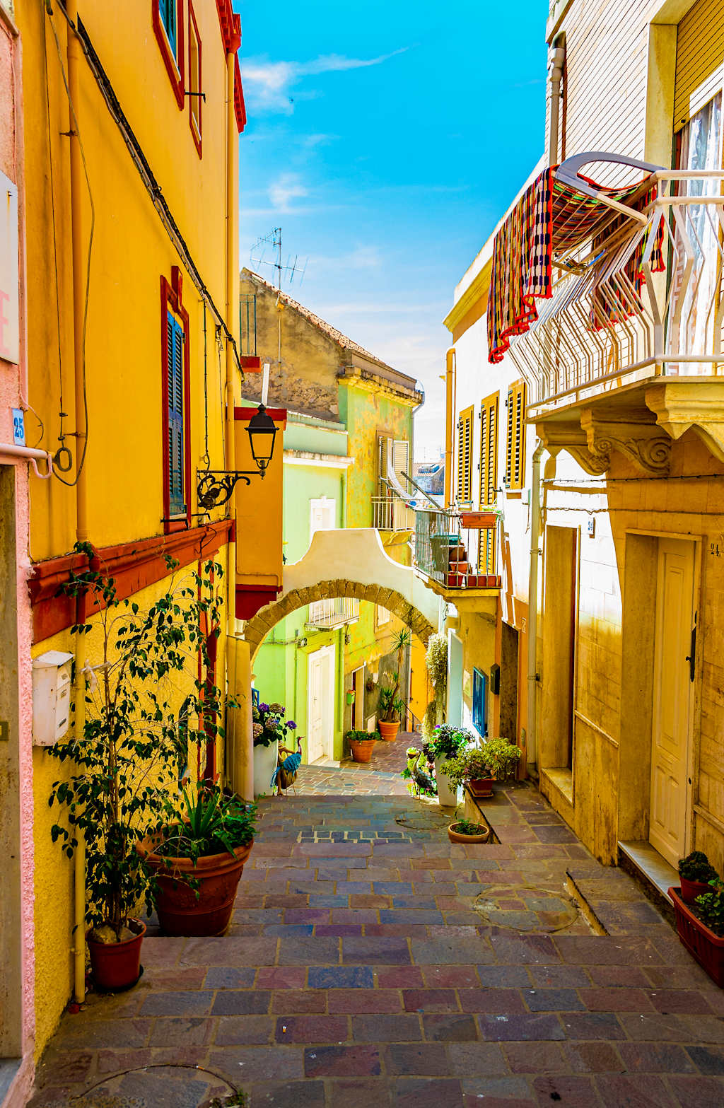 A charming alleyway lined with colorful buildings, potted plants, and a stone archway leading to a sunny courtyard in the background.