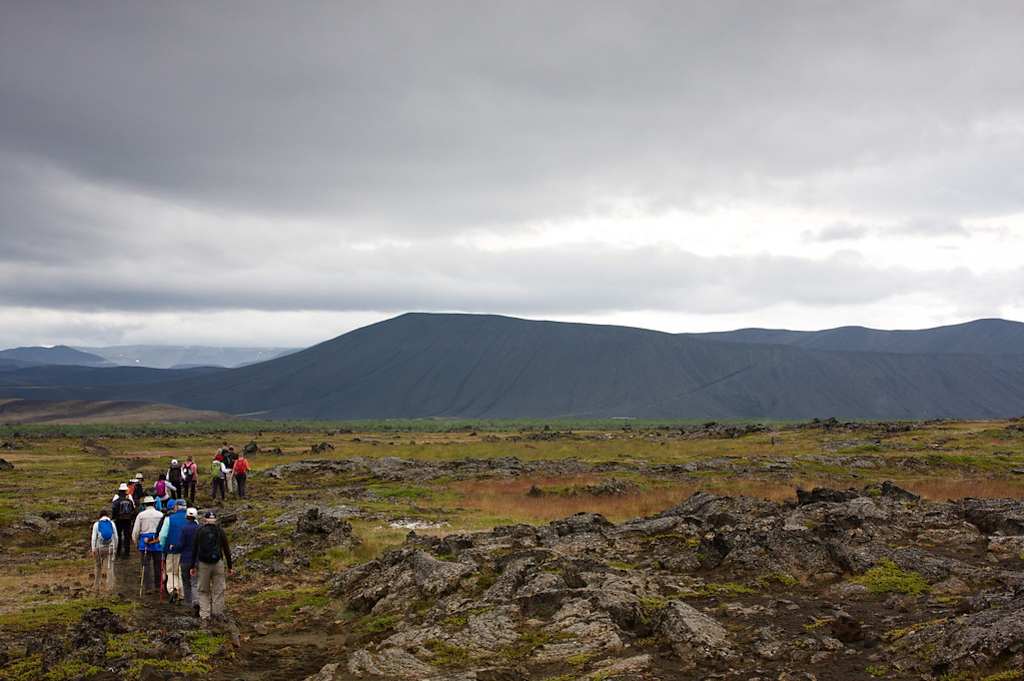 A group of people hiking through a rugged, rocky landscape with a large, imposing mountain in the background under a cloudy sky.