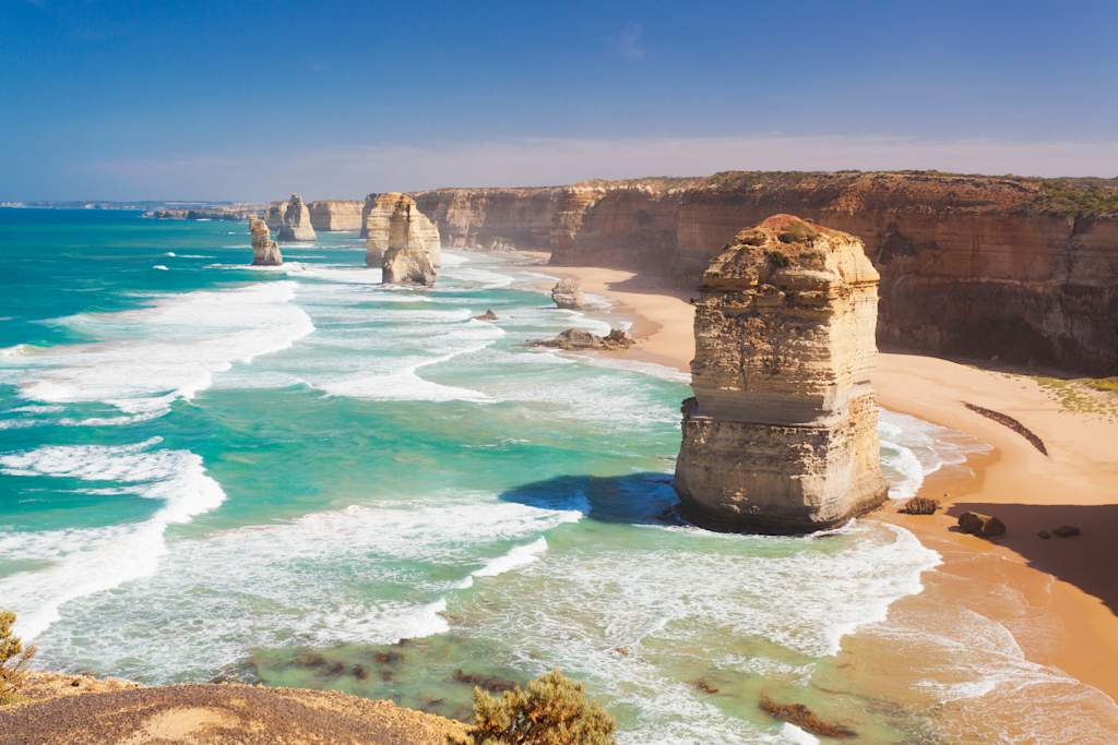 Rugged cliffs and rock formations rise from the turquoise waters, with waves crashing against the shore in the foreground.