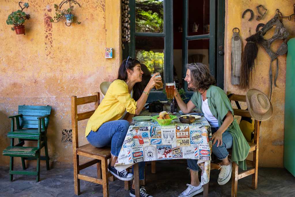 Two women sitting at a table in a rustic outdoor setting, surrounded by colorful decor and a lush, natural backdrop.