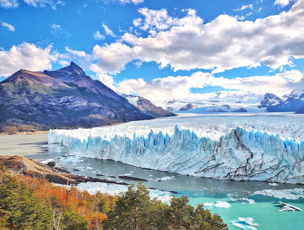 A majestic glacier with towering peaks in the background, surrounded by a serene turquoise lake and vibrant autumn foliage in the foreground.