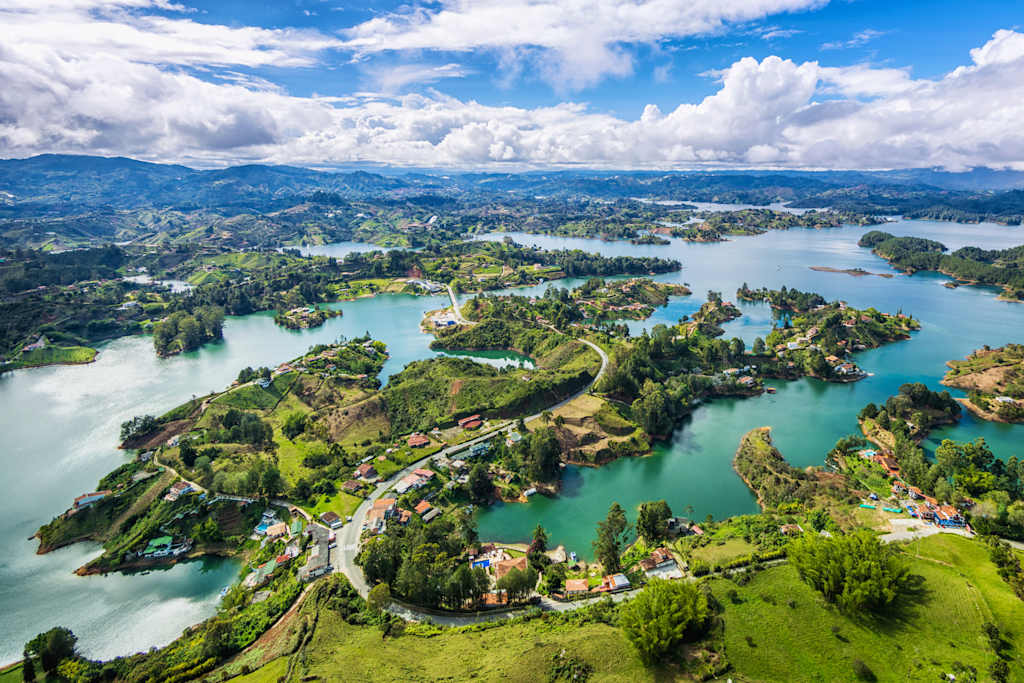 A breathtaking aerial view of a lush, green landscape dotted with islands and surrounded by crystal-clear, turquoise waters, all set against a backdrop of towering mountains and a vast, blue sky with fluffy white clouds.