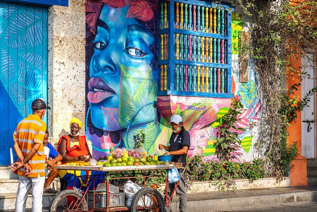 A vibrant and colorful mural adorns the wall, depicting a striking blue face with expressive features. In the foreground, a vendor sells fresh produce from a bicycle cart, adding to the lively and bustling atmosphere of the scene.