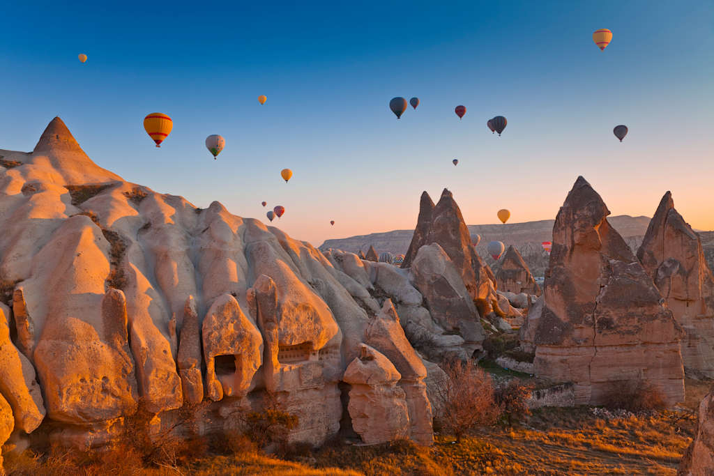 Colorful hot air balloons float above the unique rock formations of Cappadocia, Turkey, set against a vibrant blue sky at sunrise.
