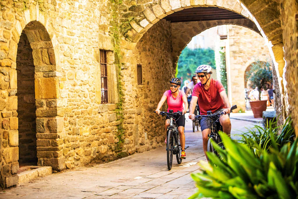 The image depicts a narrow, cobblestone alleyway flanked by stone buildings, with two cyclists in the foreground wearing helmets and brightly colored jerseys, riding through the archway.
