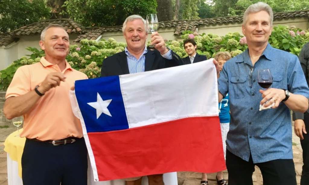 A group of four older men, dressed casually, are holding a large Chilean flag and appear to be celebrating or expressing pride in their national identity.