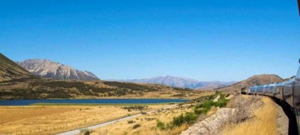 A scenic landscape with a winding dirt path along a lake, surrounded by rugged mountains under a clear blue sky.
