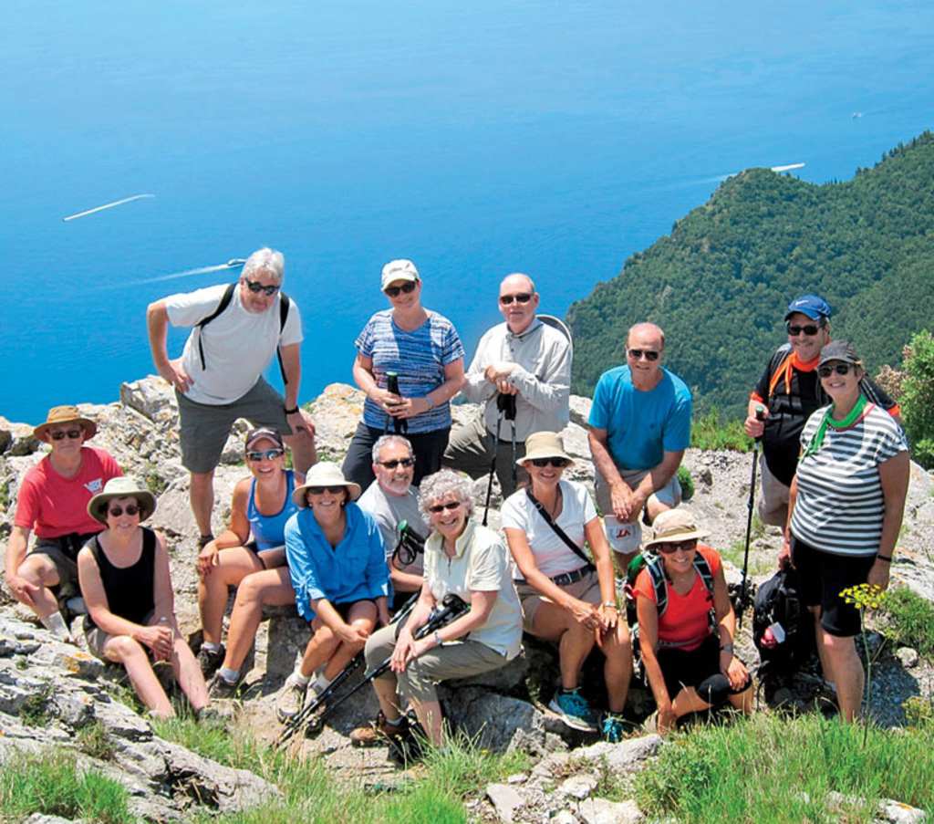 A group of hikers posing for a photo on a rocky, grassy hillside with a scenic blue ocean and forested mountains in the background.
