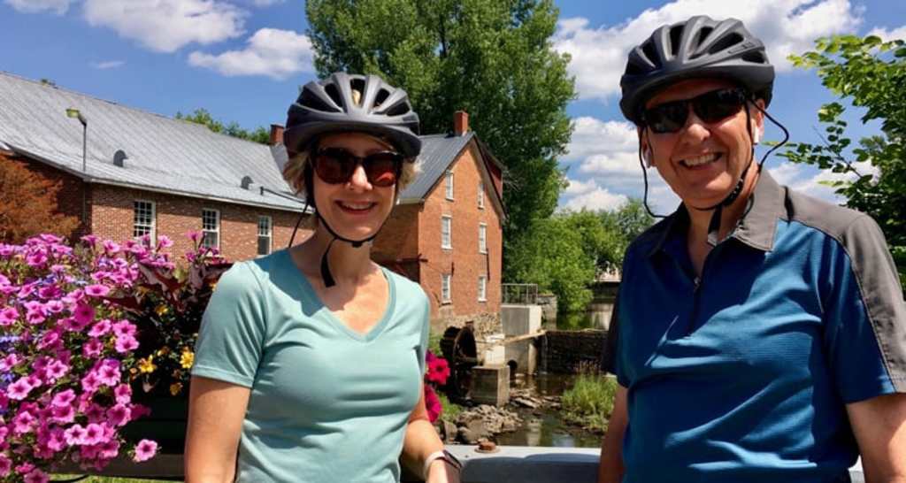 Two people wearing bicycle helmets and sunglasses stand in front of a brick building surrounded by lush greenery and flowers.
