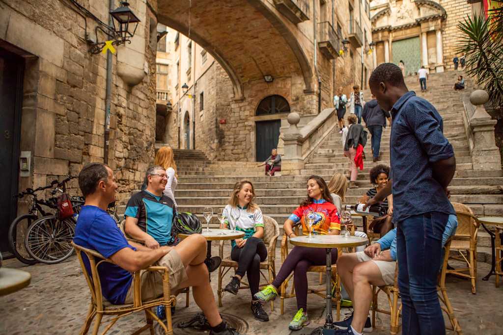 A group of people, both men and women, are sitting at outdoor tables in a historic European-style alleyway, surrounded by old stone buildings and arches.