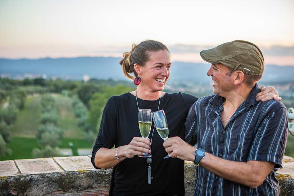 A smiling couple holding champagne flutes, with a scenic landscape of mountains and forests in the background.