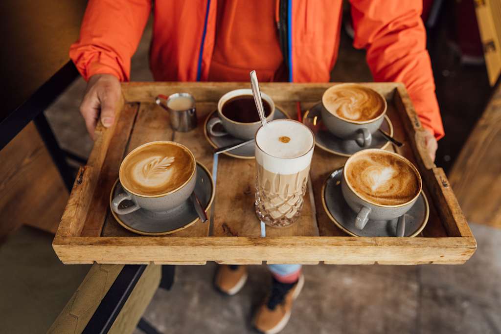 A wooden tray holds several cups of steaming coffee with intricate latte art, accompanied by a person wearing a vibrant orange jacket in the background.
