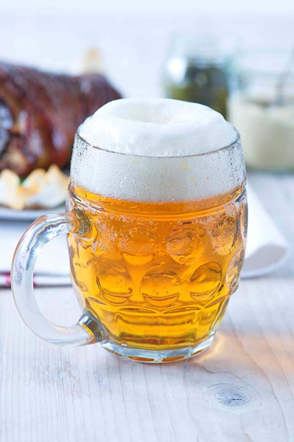A glass mug filled with a golden-colored liquid, topped with a thick layer of white foam, sits on a wooden surface alongside a plate of what appears to be barbecued or grilled meat.