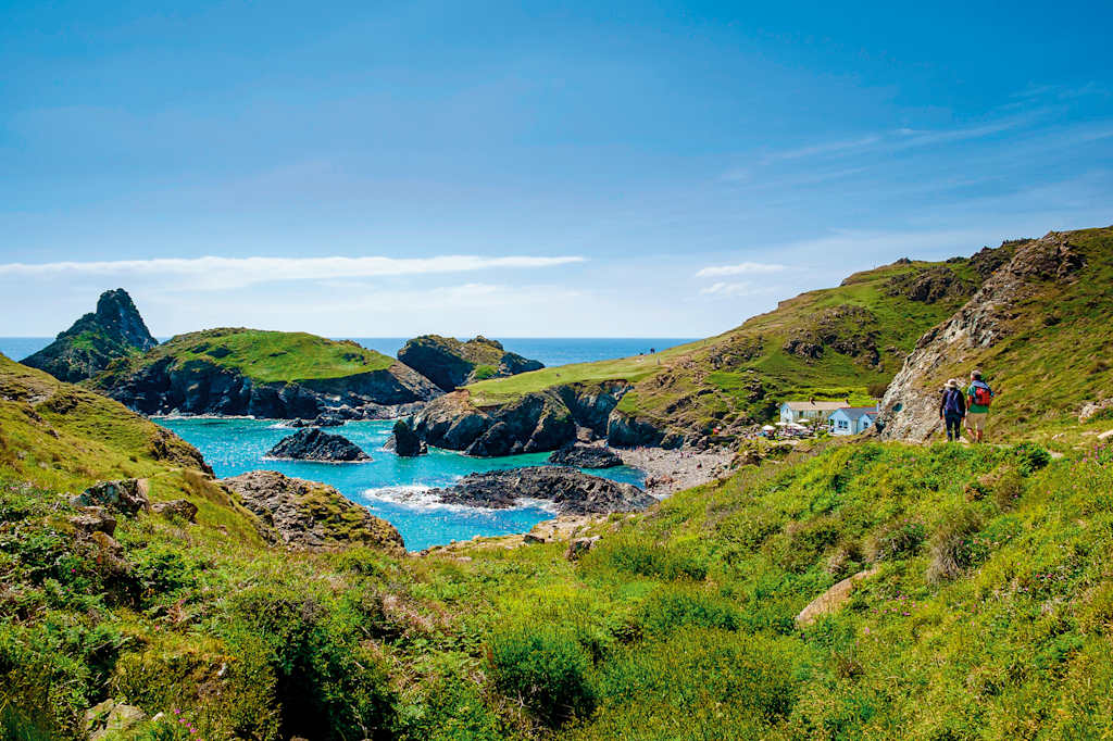 A lush, green landscape with rocky cliffs and a vibrant turquoise bay surrounded by towering peaks in the distance, with a person hiking along the trail in the foreground.
