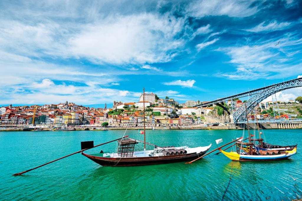 A picturesque view of the historic city of Porto, Portugal, with its colorful buildings, iconic Dom Luís I Bridge, and traditional boats known as rabelos floating on the serene waters of the Douro River.