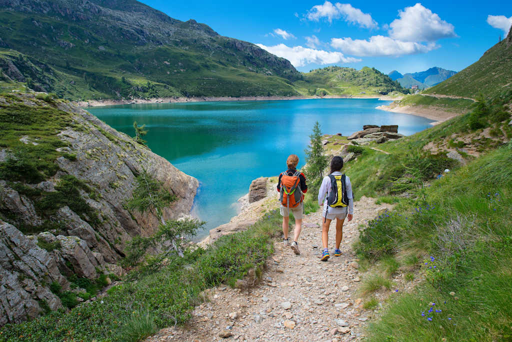 Two hikers walking on a dirt path along a stunning turquoise lake surrounded by rugged mountains and lush greenery under a bright blue sky with fluffy white clouds.