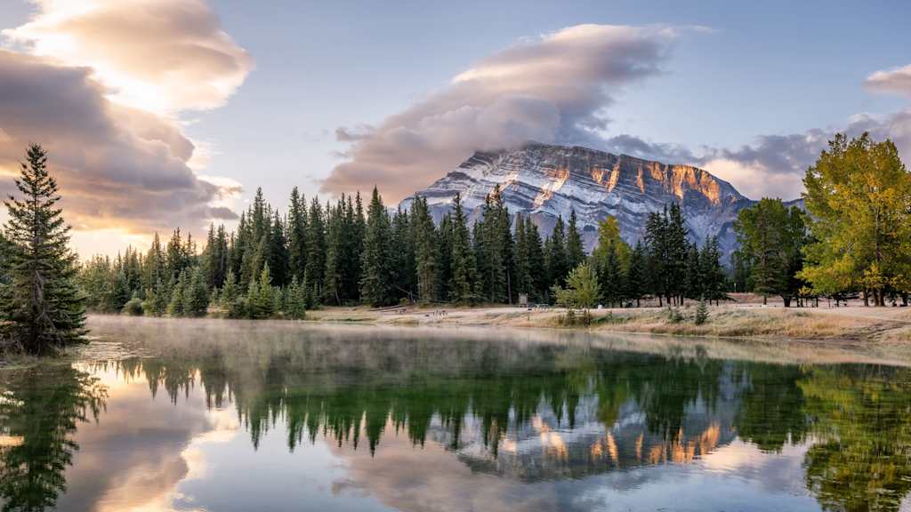 A serene lake reflects the majestic snow-capped mountain surrounded by a dense forest, creating a breathtaking landscape under a dramatic cloudy sky.