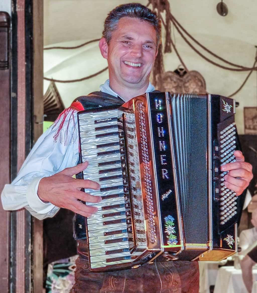 A smiling man holding a large accordion-like instrument, surrounded by various items in what appears to be a rustic or rural setting.