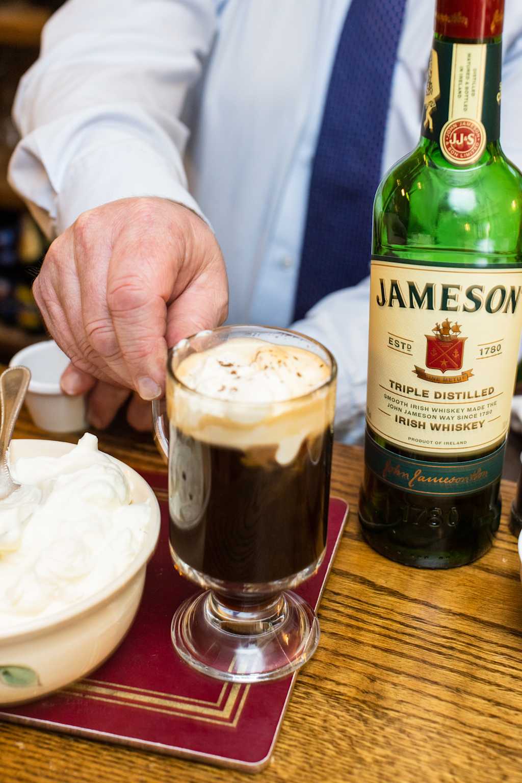 A person's hands holding a glass of coffee or hot chocolate, with a bottle of Jameson Irish whiskey visible in the background on a wooden table.