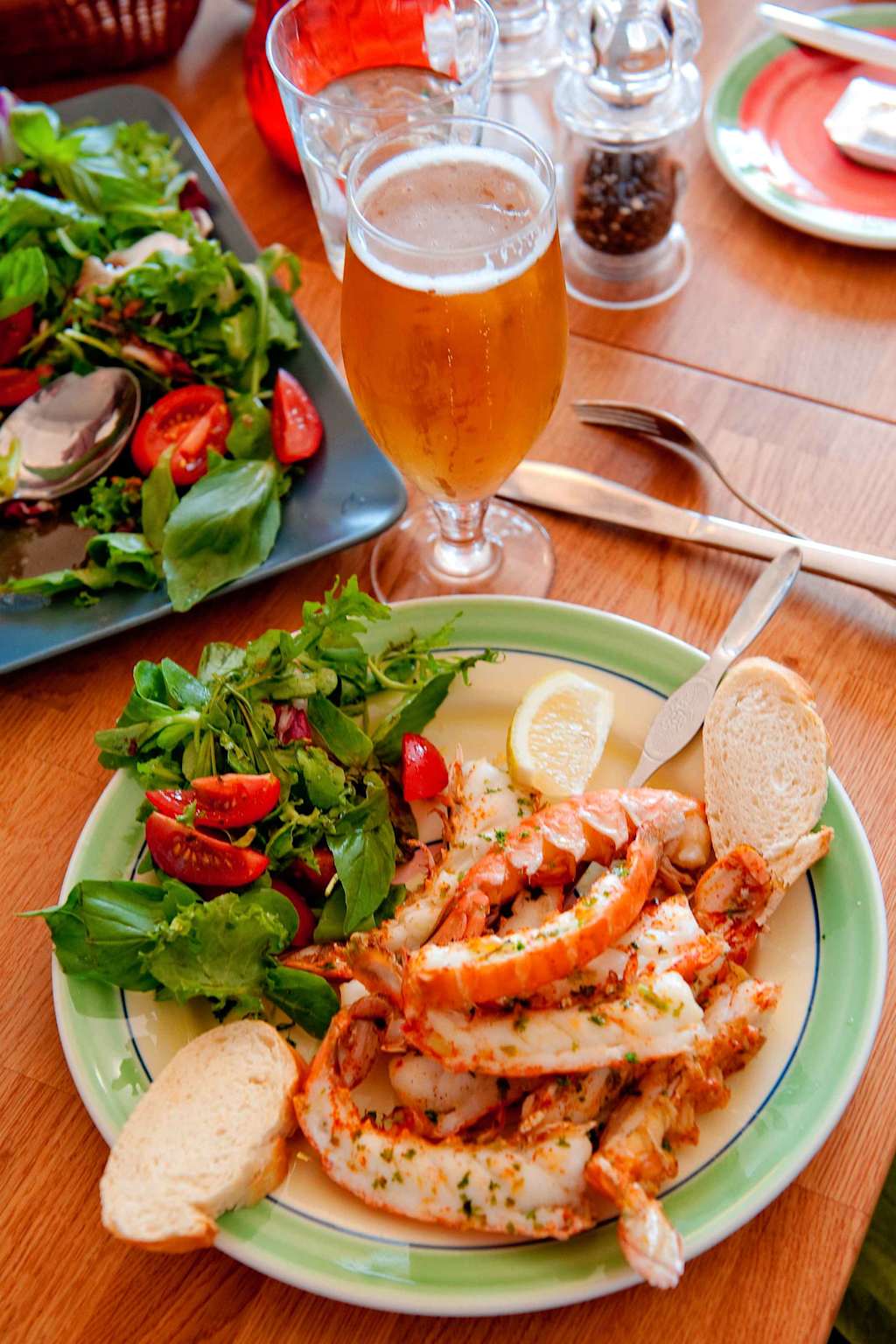 A plate of grilled fish or seafood, accompanied by a fresh salad and a glass of amber-colored beverage, sits on a wooden table alongside other dining items.