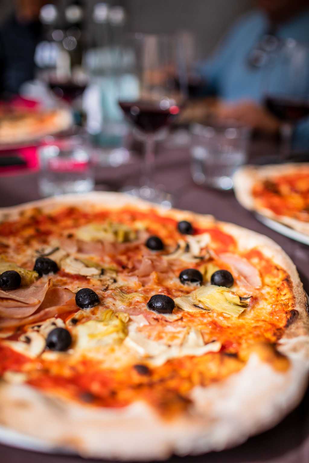 A delicious-looking pizza with various toppings, including olives and ham, sits on a table in a restaurant setting with blurred background elements.