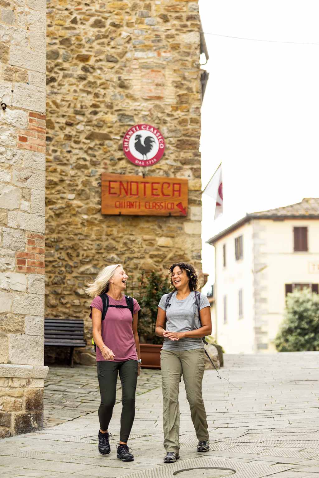 Two women are walking and conversing in a town square, with a stone building and a sign for "ENOTECA" visible in the background.