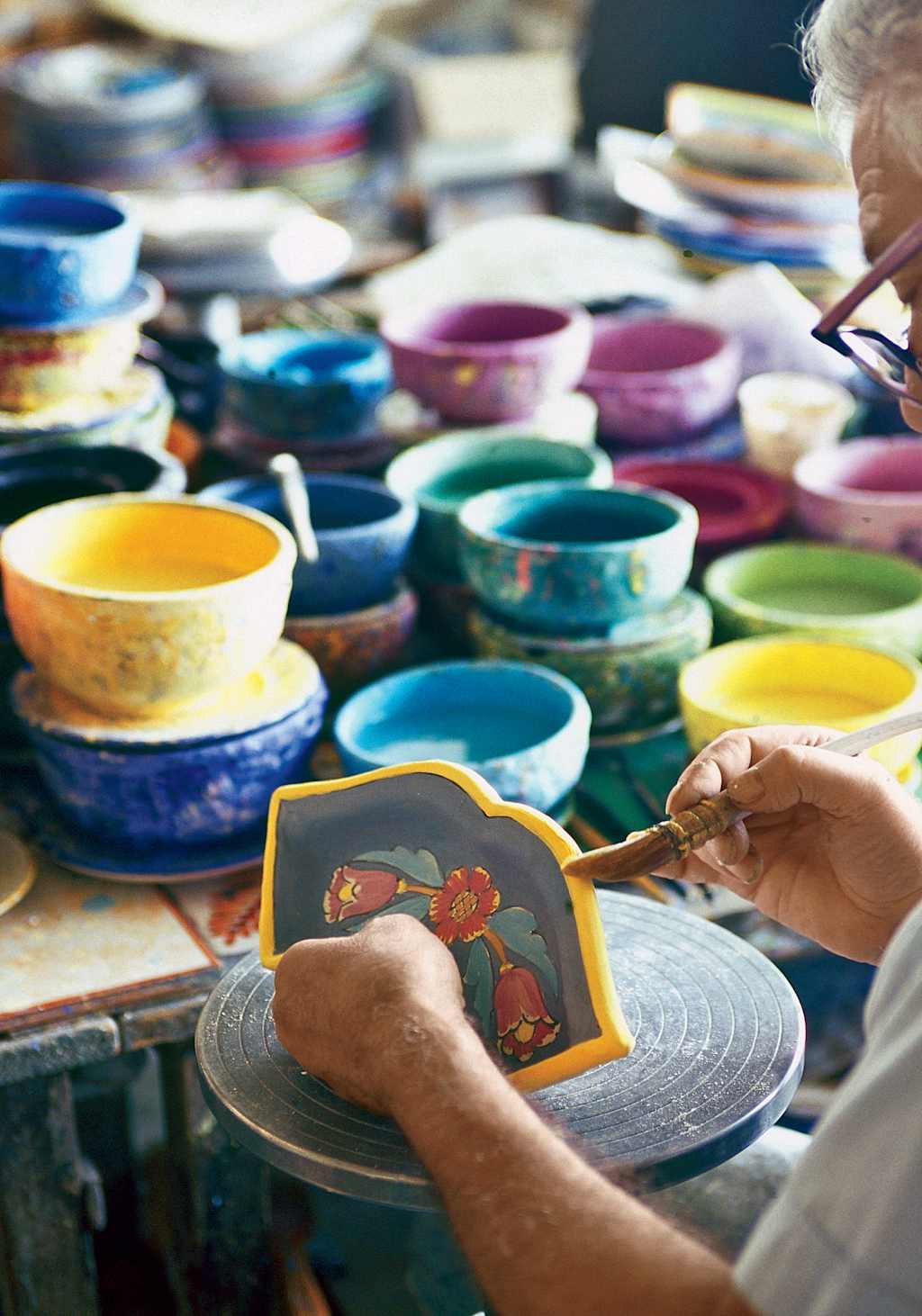 A variety of colorful ceramic bowls and plates are displayed on a table, with a person's hands working on decorating one of the ceramic pieces.