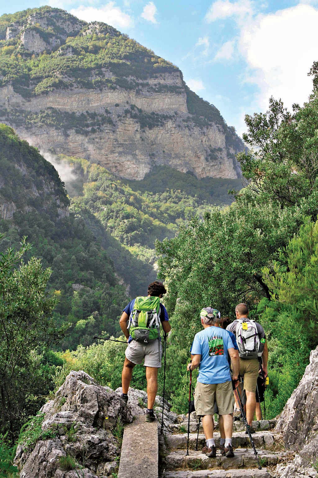 A group of hikers traversing a rocky trail through a lush, mountainous landscape with towering cliffs in the background.