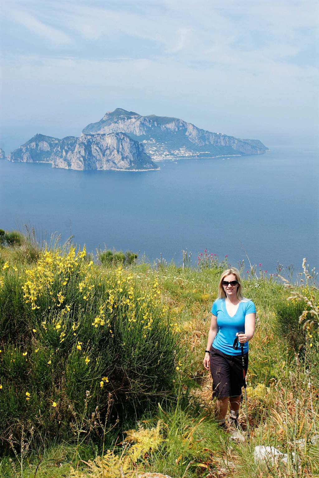 A woman in a blue shirt and sunglasses walks along a trail surrounded by yellow wildflowers, with a scenic coastal landscape featuring rocky cliffs and the sea visible in the background.