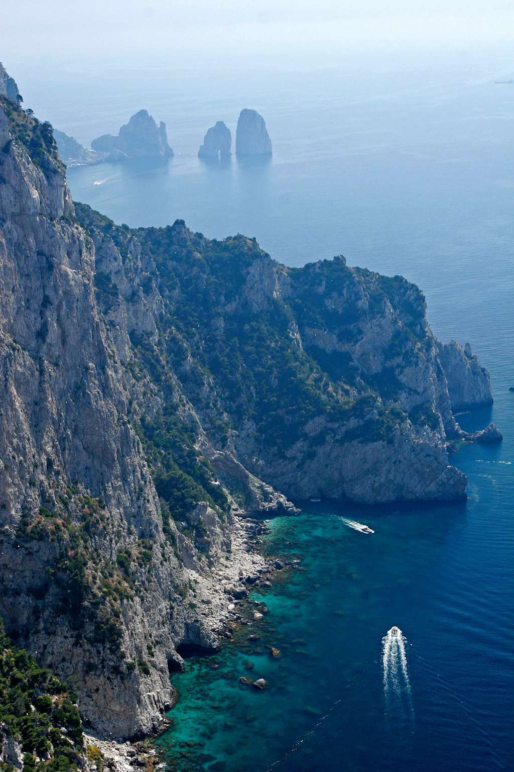 A rugged, mountainous coastline with towering cliffs overlooking a serene, turquoise sea dotted with small islands in the distance.