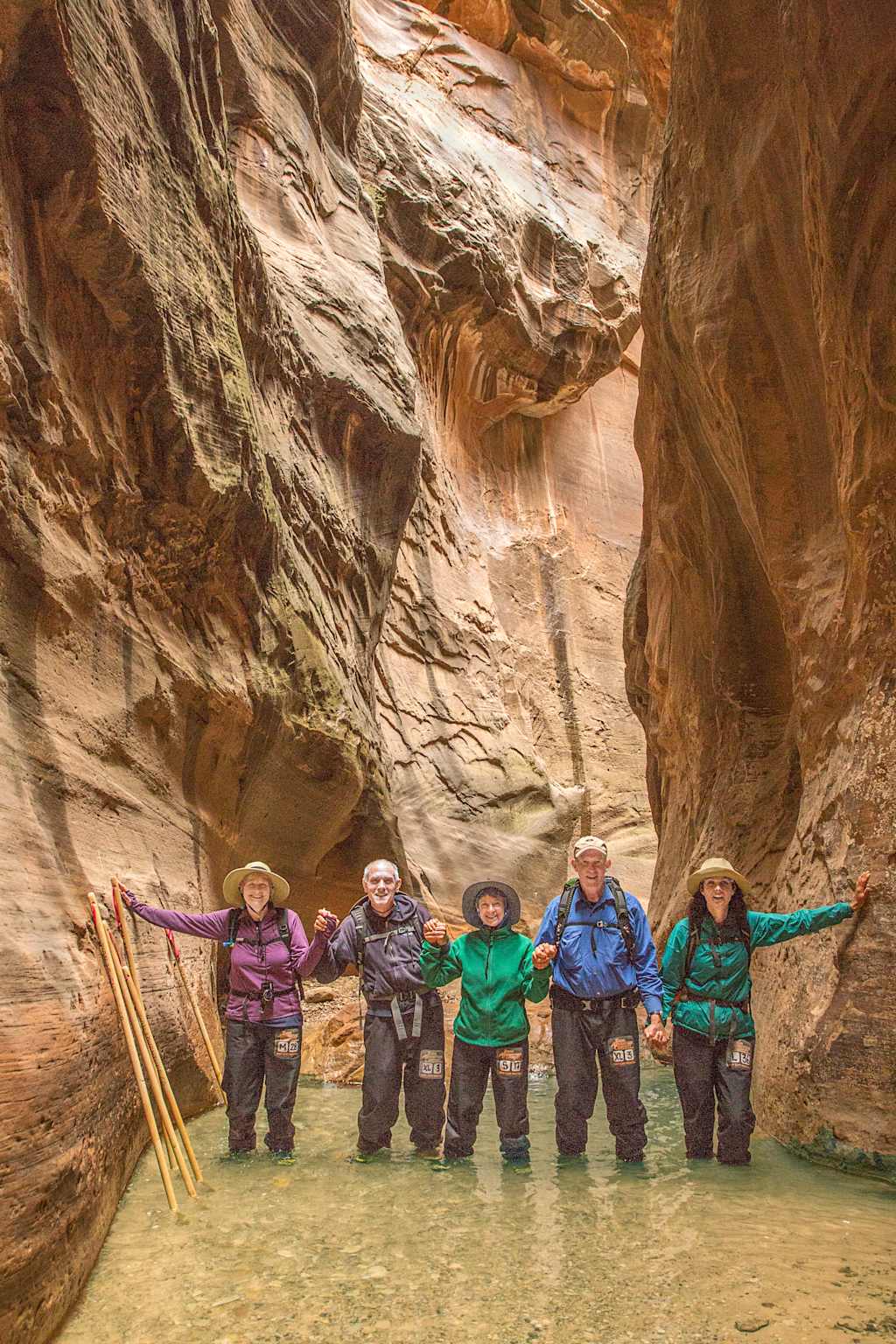 A group of hikers standing in a shallow stream surrounded by towering, multicolored rock formations in a narrow canyon.
