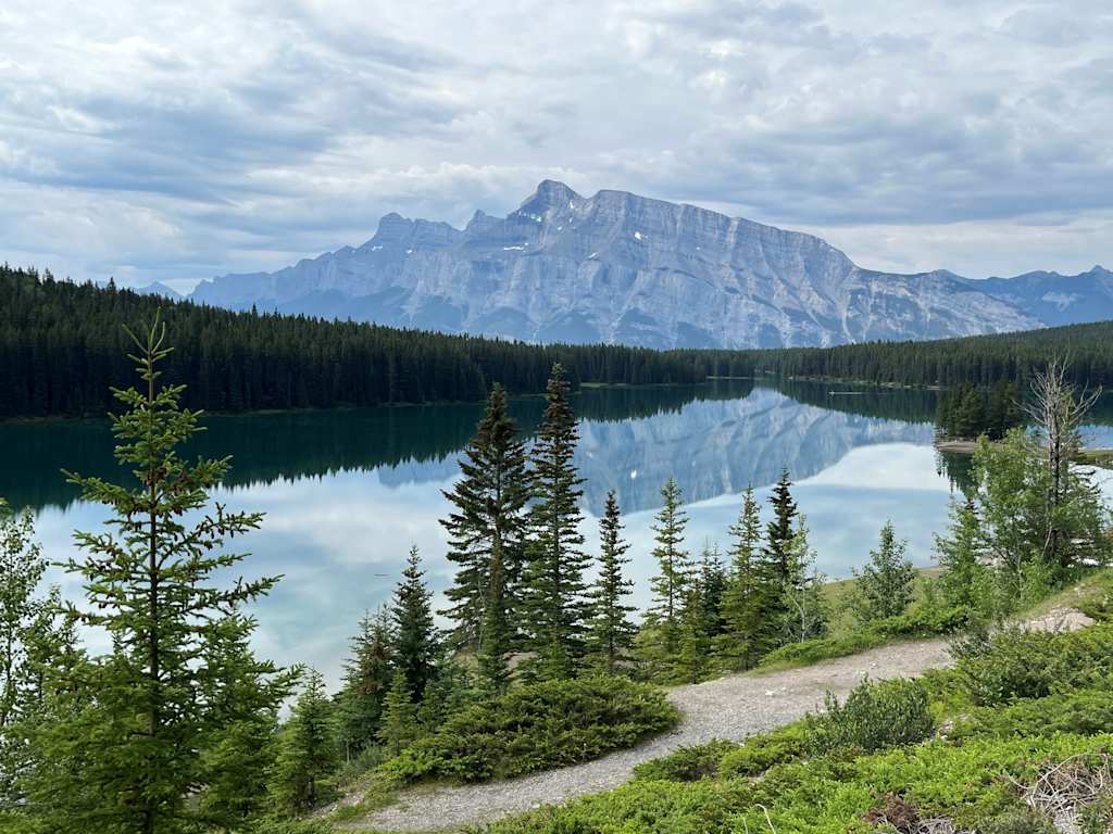 A serene mountain lake surrounded by lush evergreen forests and towering snow-capped peaks under a dramatic cloudy sky.
