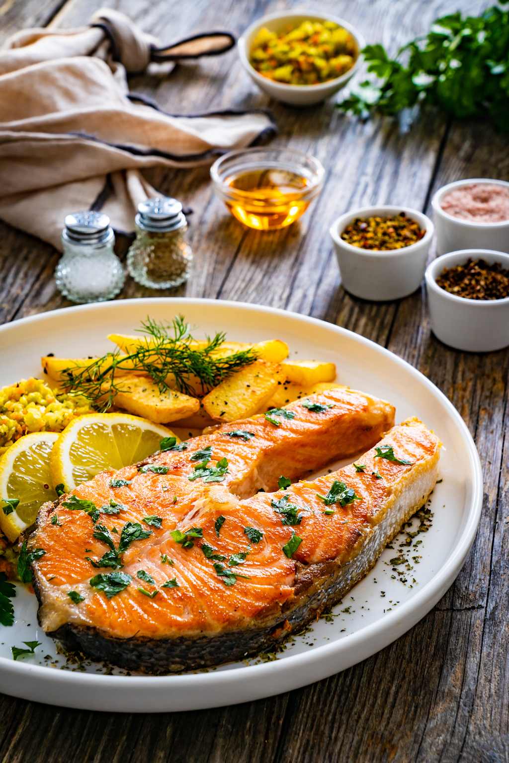 A plate of grilled salmon fillet served with lemon slices, herbs, and roasted potatoes on a wooden table, surrounded by various spices and condiments.