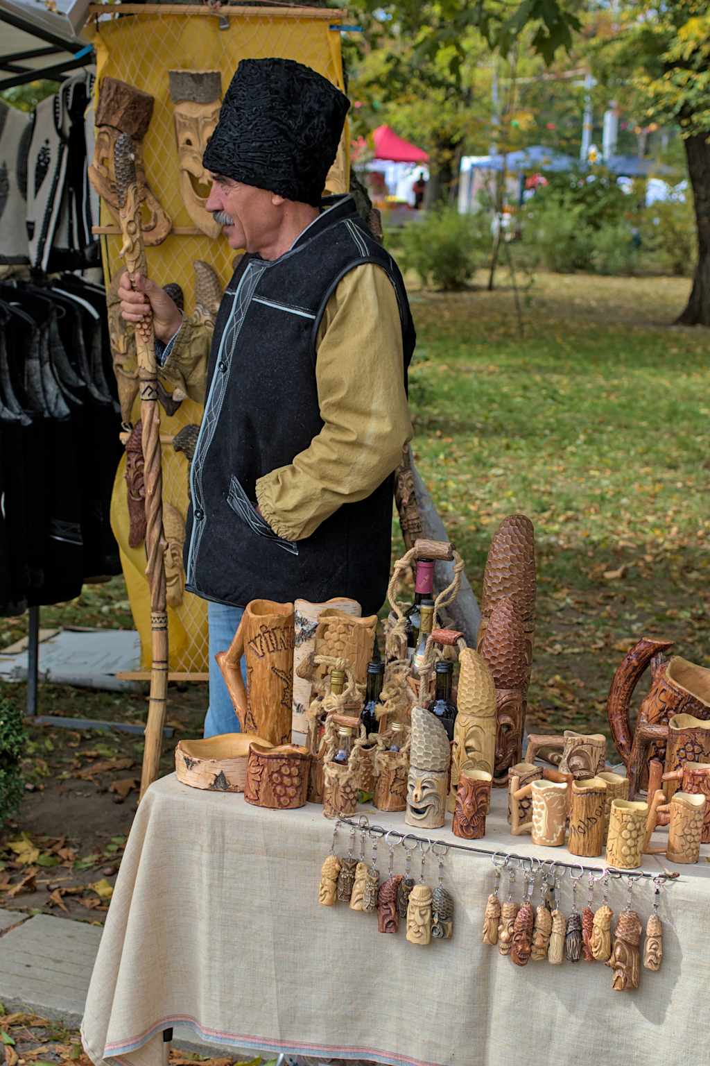 A person in a black hat and vest is selling various handcrafted wooden items on a table in an outdoor setting with trees and other vendors in the background.