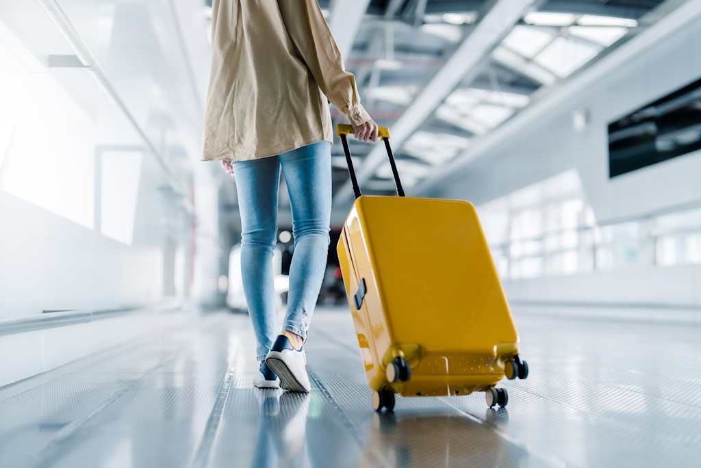 A person in casual attire, carrying a yellow suitcase, walking through a modern, well-lit airport terminal.
