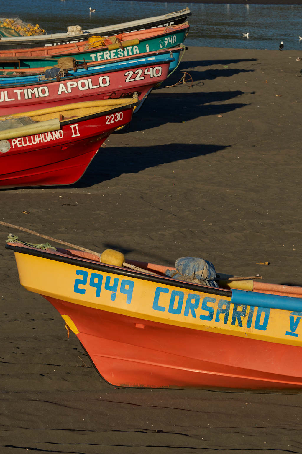 Colorful fishing boats with various names and numbers are docked in the harbor, with a calm body of water and a cloudy sky in the background.