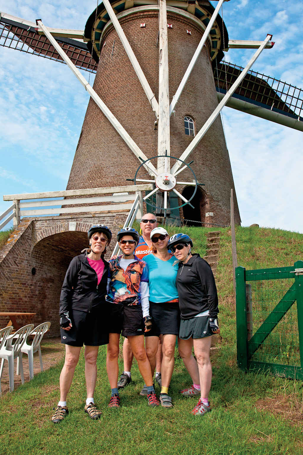 A group of people standing in front of a large, traditional windmill in a rural setting, with a clear blue sky in the background.