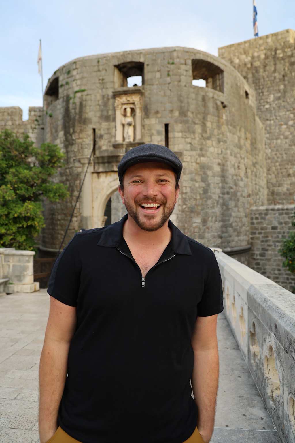 A smiling man wearing a black shirt and cap stands in front of a large stone castle or fortress in the background.
