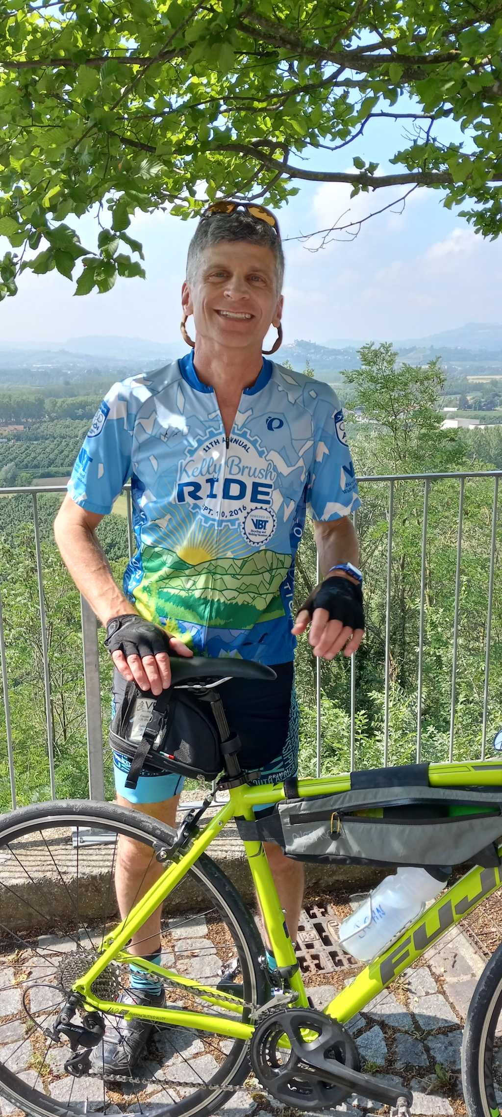 A smiling older man in a cycling jersey stands next to a brightly colored bicycle, with a scenic landscape of trees and hills visible in the background.