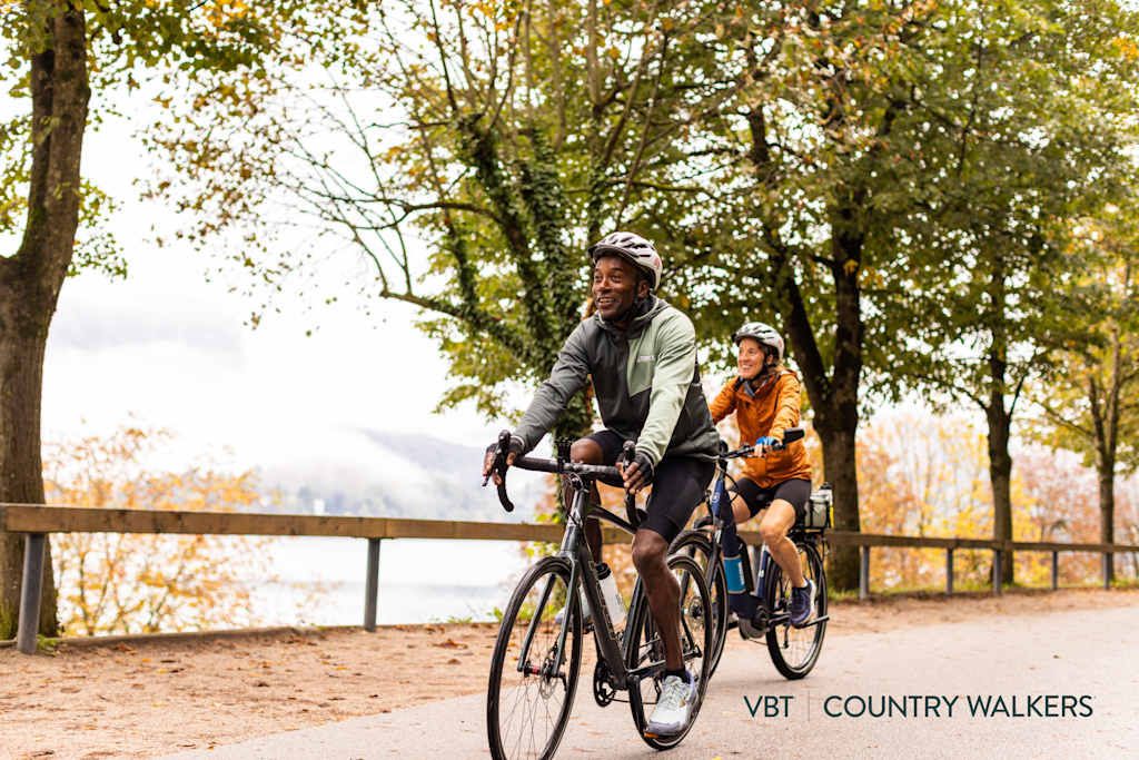 Two individuals, a man and a woman, are riding bicycles on a path surrounded by lush, autumn-colored trees. The foreground features the cyclists, while the background showcases the scenic, natural environment.