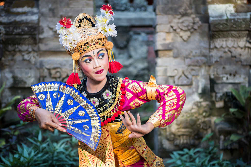 A colorfully dressed performer with an elaborate headdress and fan poses in front of a stone wall backdrop, surrounded by lush greenery.