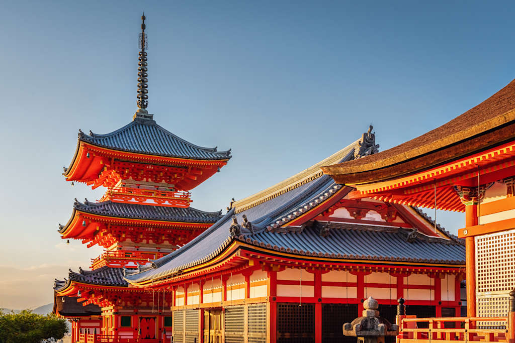 A traditional Japanese pagoda with its distinctive red and gold architecture stands prominently in the foreground, set against a clear blue sky in the background.