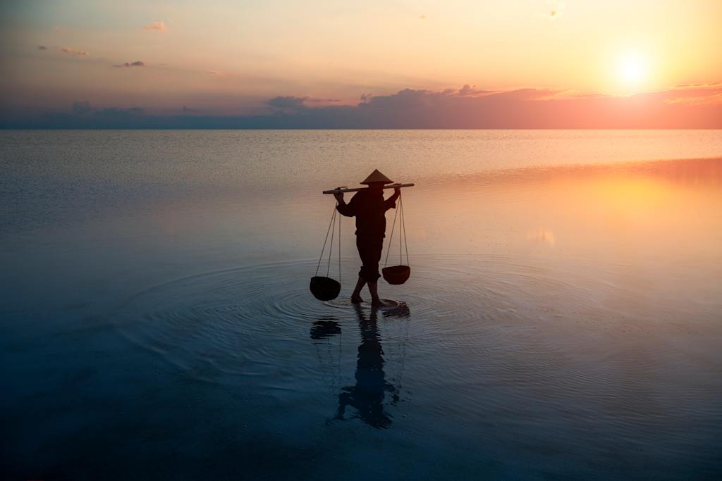 A silhouetted figure stands on a calm body of water, carrying baskets on a pole, against a serene sunset backdrop with clouds and reflections.