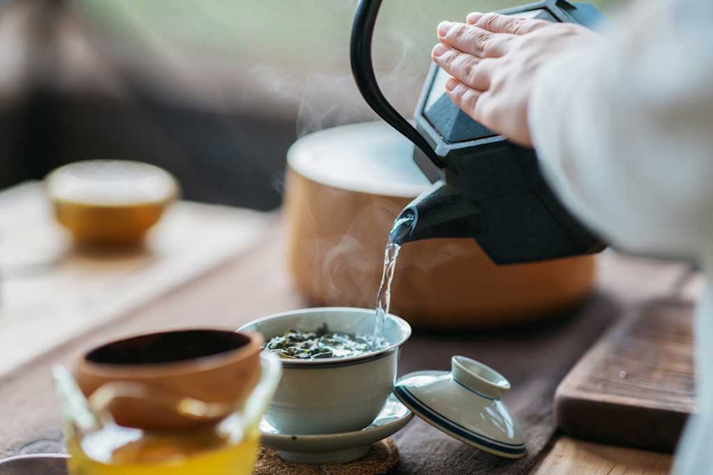 A person's hands are pouring hot water from a kettle into a ceramic cup, with a wooden table and other objects in the background.