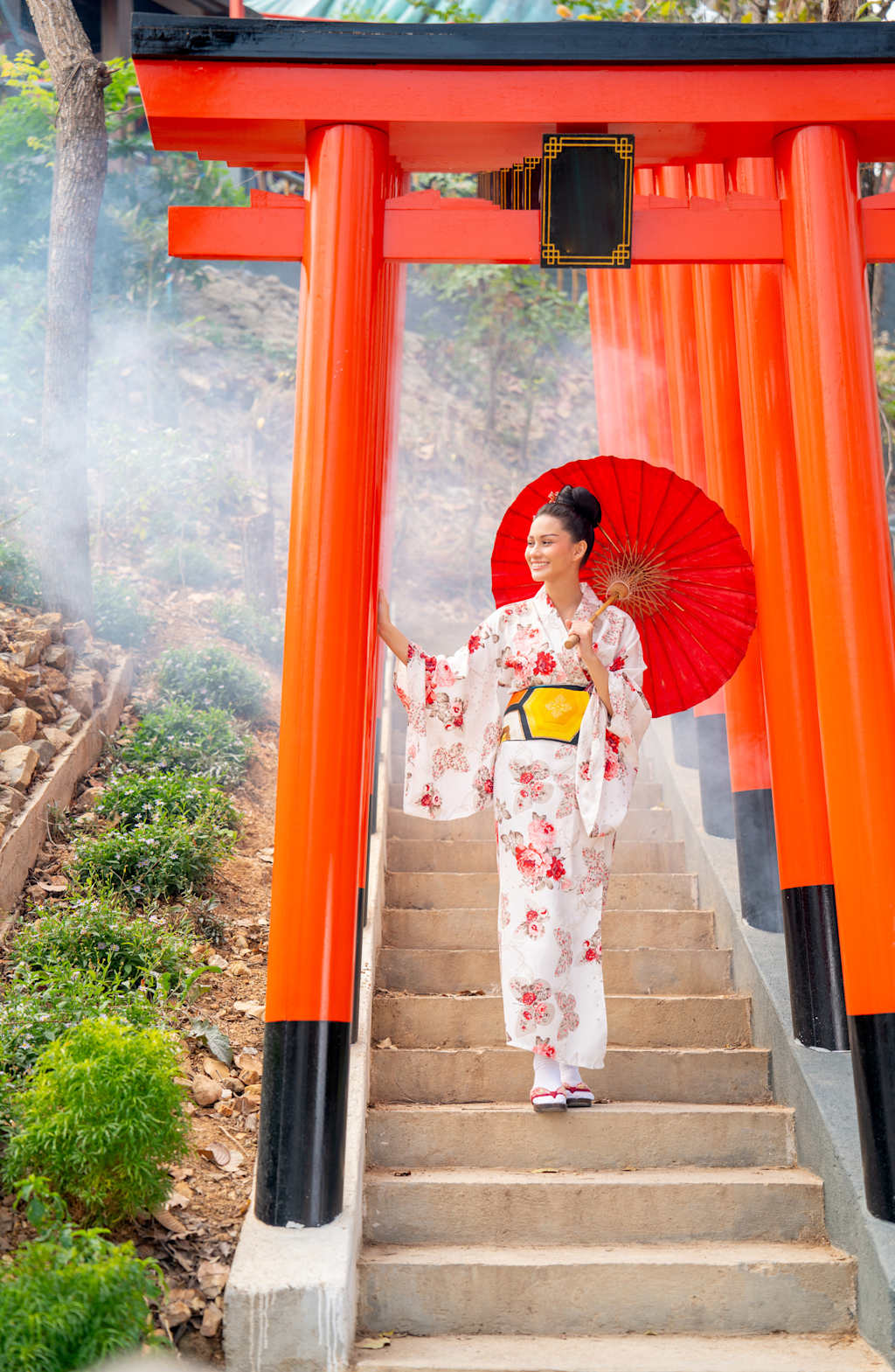 A woman in a traditional Japanese kimono with a red umbrella stands on the steps of a vibrant red Torii gate, surrounded by lush greenery.