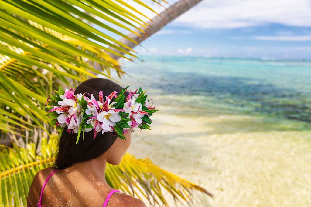 A person wearing a flower crown stands on a beach, with palm fronds framing the view of the turquoise ocean in the background.